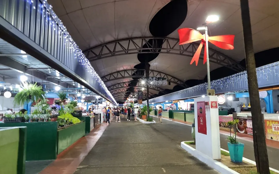 Corredor coberto da Feira Central de Campo Grande à noite, com bancas iluminadas, plantas e visitantes