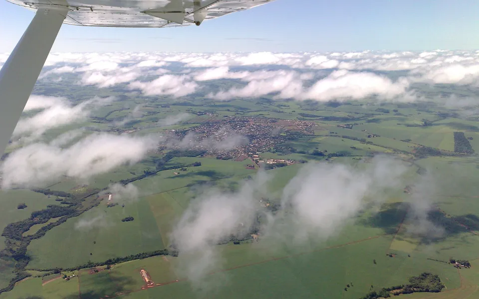 Vista aérea de Ubiratã, no Paraná, entre lavouras verdes, com nuvens baixas e a asa de um avião em primeiro plano