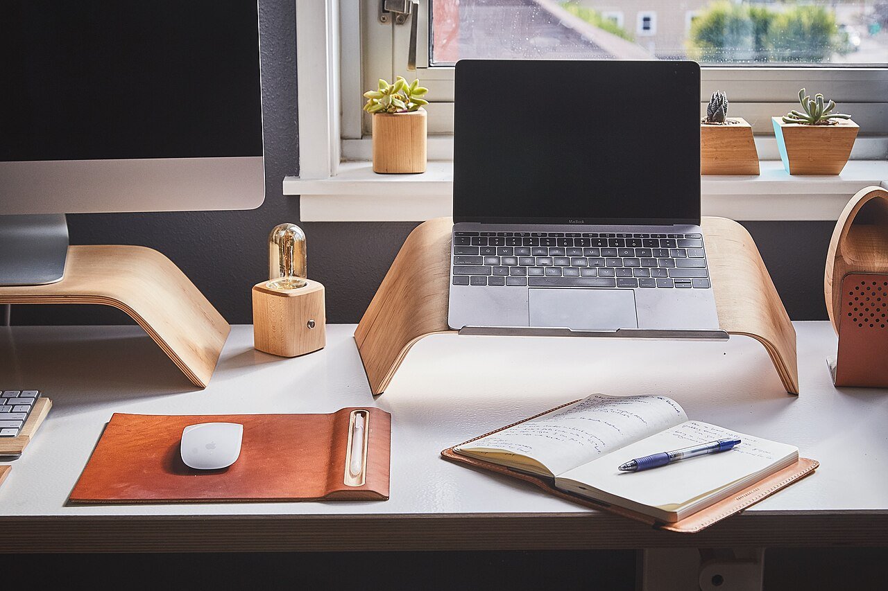 Mesa de trabalho com computador, notebook e um caderno aberto com caneta